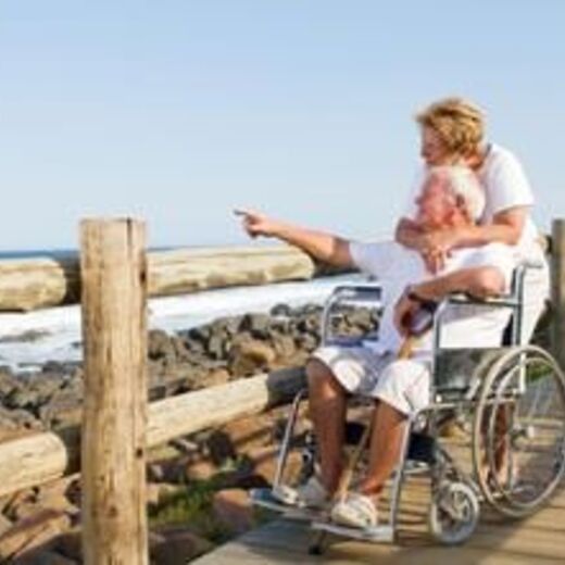 Senior im Rollstuhl zeigt auf das Meer während seine Frau ihn umarmt
