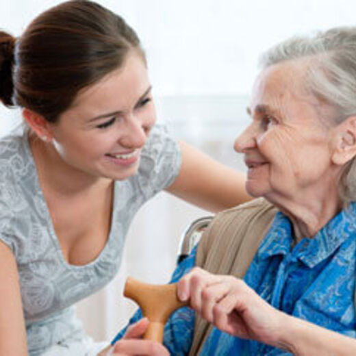 Ein junge Frau lächelt eine Seniorin mit Gehstock an