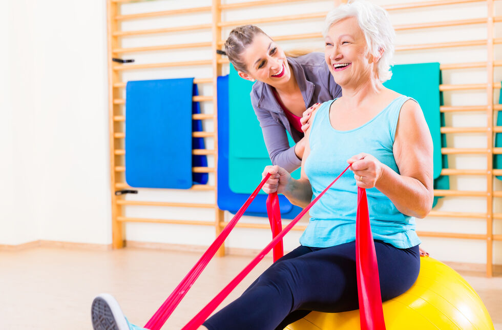 Seniorin turnt auf einem Gymnastikball mit Dehnband.