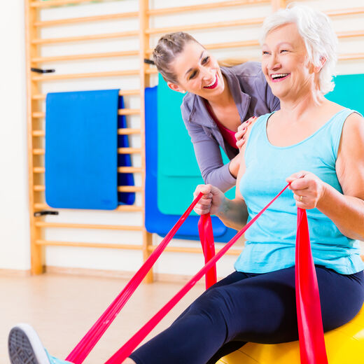 Senioren auf Gymnastikball macht Übungen mit Fitnessband unter Betreuung