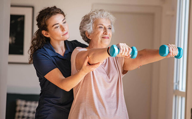 Seniorin trainiert mit Hanteln ihre Armmuskulatur und wird von Physiotherapeutin unterstützt. © Rido - stock.adobe.com