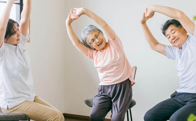 Seniorenpaar führt gemeinsam mit Therapeutin Gymnastikübungen im Sitzen durch. © buritora - stock.adobe.com