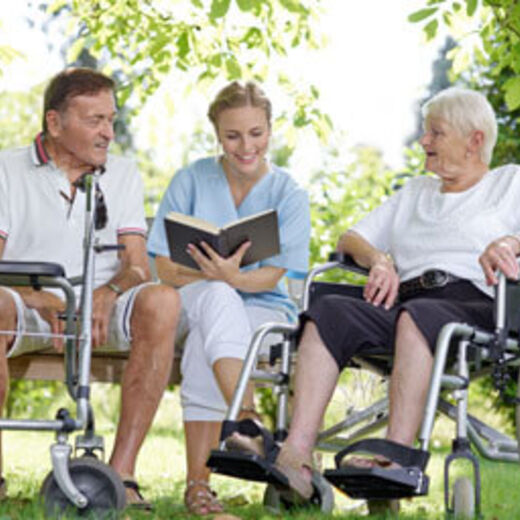 Ein Mann mit Rollator, eine Frau mit einem Buch in der Hand und eine Frau im Rollstuhl sitzen im Park