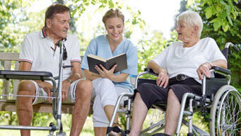 Ein Mann mit Rollator, eine Frau mit einem Buch in der Hand und eine Frau im Rollstuhl sitzen im Park