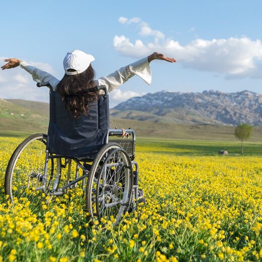 Portraits: Eine Frau im Rollstuhl steht mitten auf einer Wiese und streckt die Arme in die Luft
