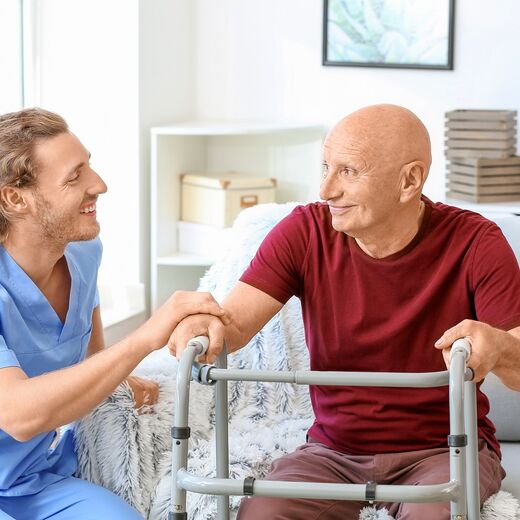 Pfleger hilft einem älteren Mann mit Rollator von dem Sofa aufzustehen