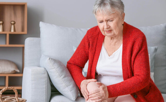 Seniorin sitzt auf der Couch und hält Ihr Knie fest, weil es schmerzt. © Pixel-Shot - stock.adobe.com