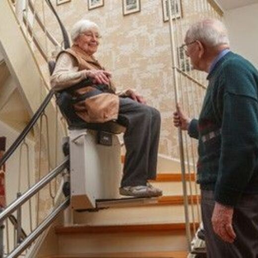 Senorin fahr auf dem Sitzlift die Treppen hoch während ihr Mann zuschaut