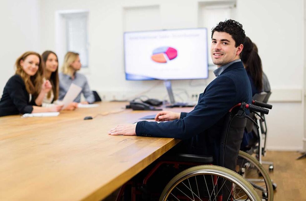 Leben mit Behinderung: Ein Mann im Rollstuhl sitzt mit Kolleg:innen im Meeting-Raum