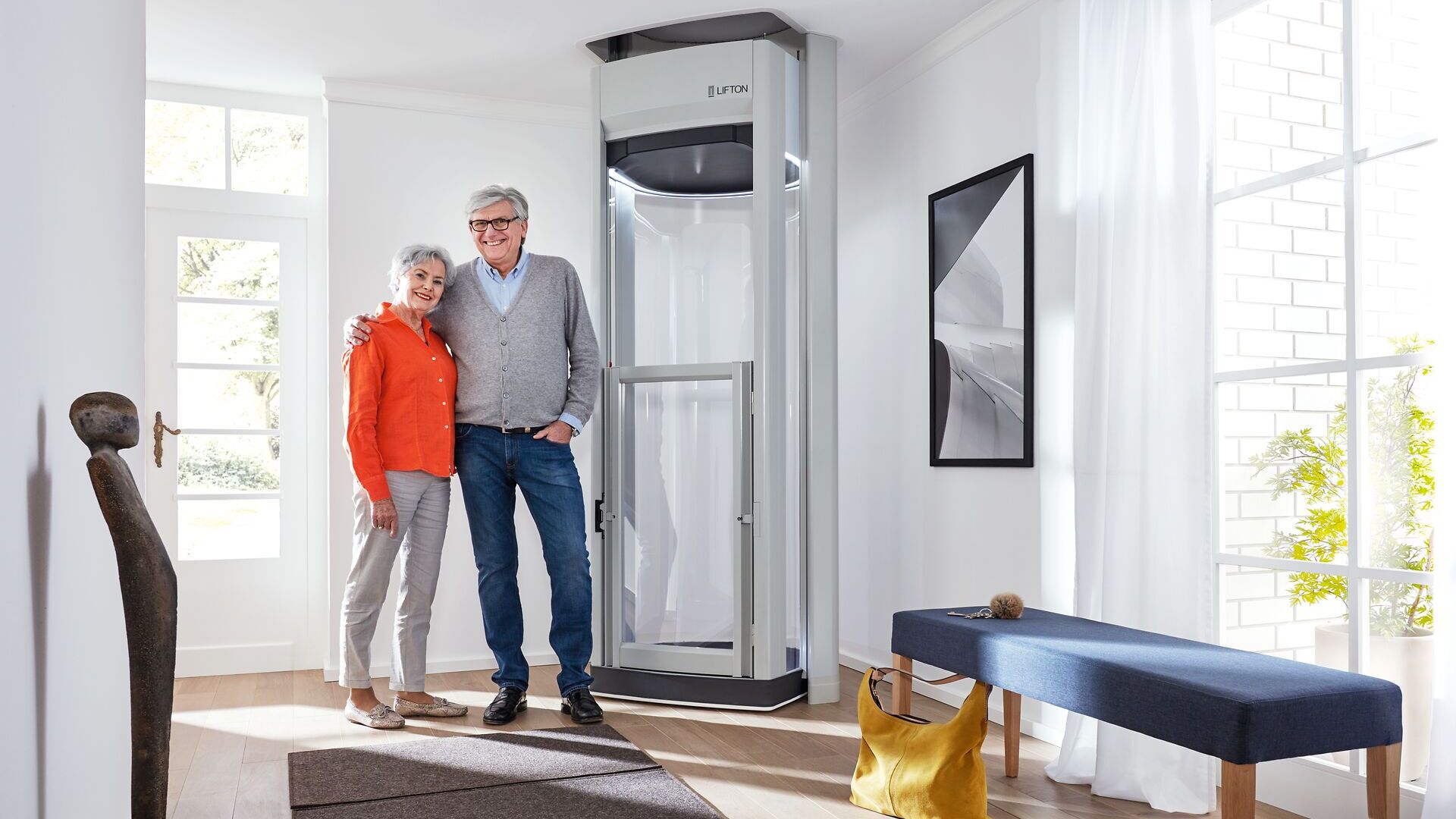 Älteres Paar steht lächelnd neben einem modernen Treppenlift im Wohnzimmer barrierefreies Wohnen Treppenlift-Ratgeber.